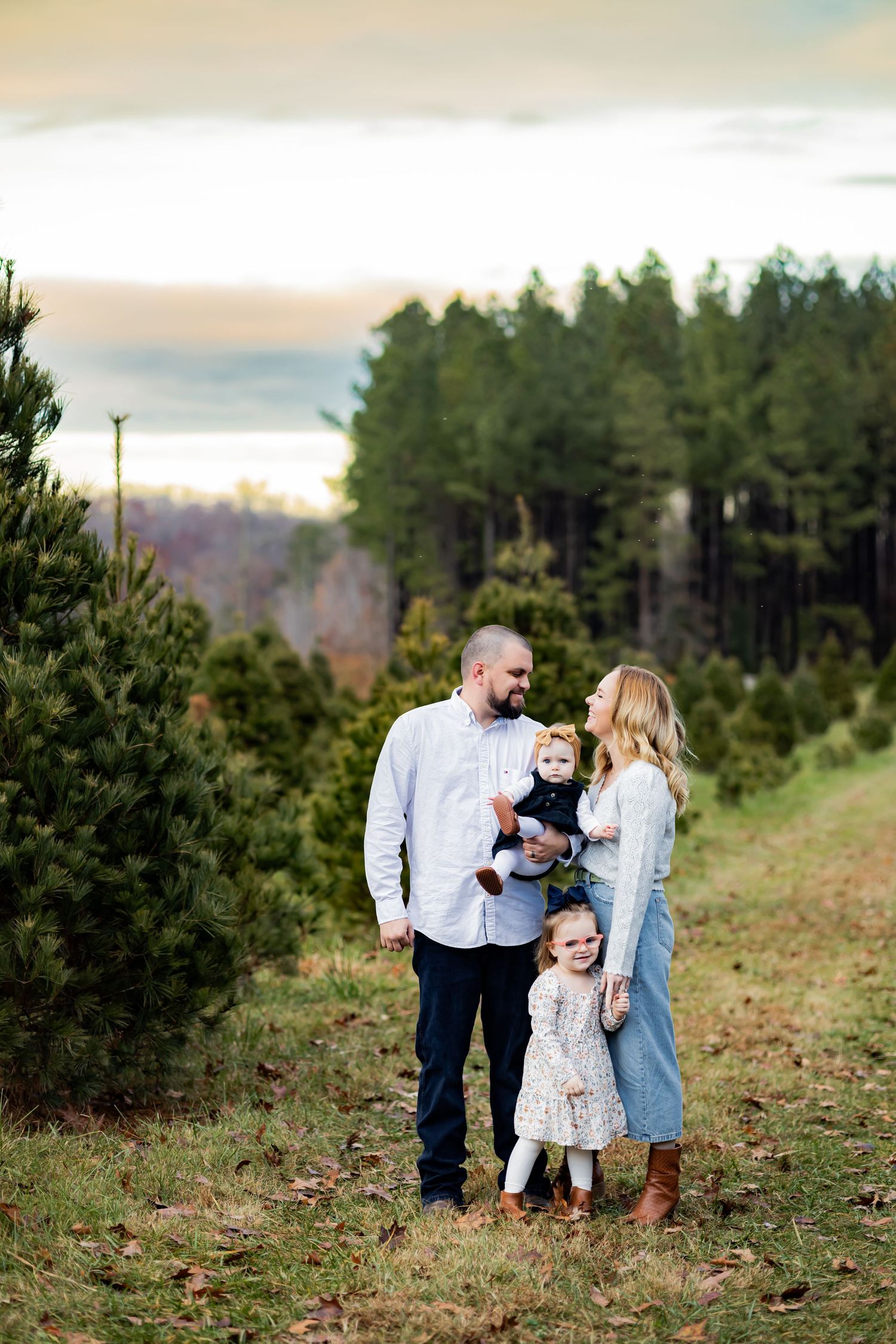A family enjoys a tender moment together on a grassy hillside with evergreen trees in the background. RVA Family photographer. Photographer