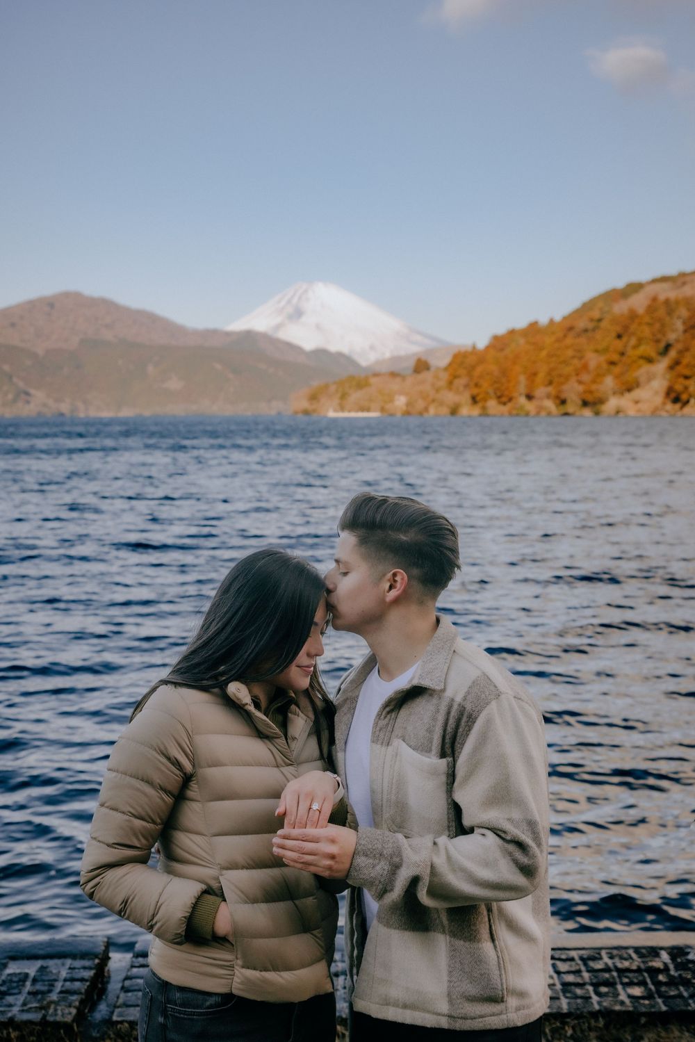 A couple shares a romantic moment by Lake Ashi with Mount Fuji visible in the background on an autumn day.