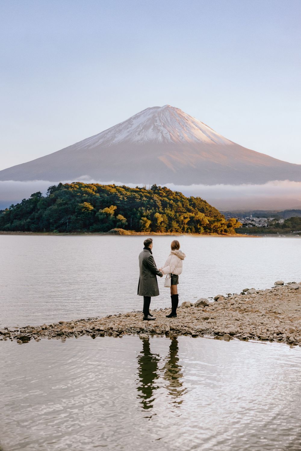 Two figures stand on a rocky shore with Mount Fuji rising majestically through misty clouds in the background.