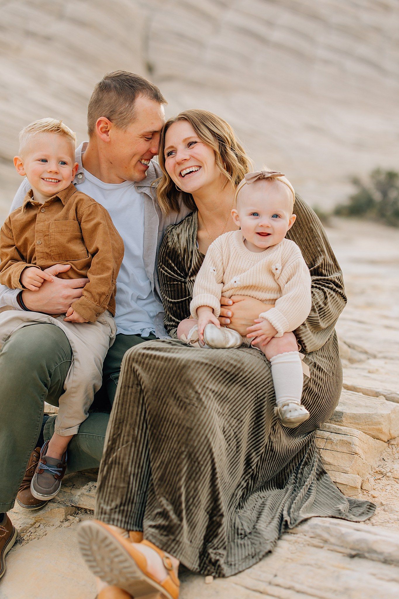 A happy family of four sits together on sandy rocks during a warm outdoor photoshoot.