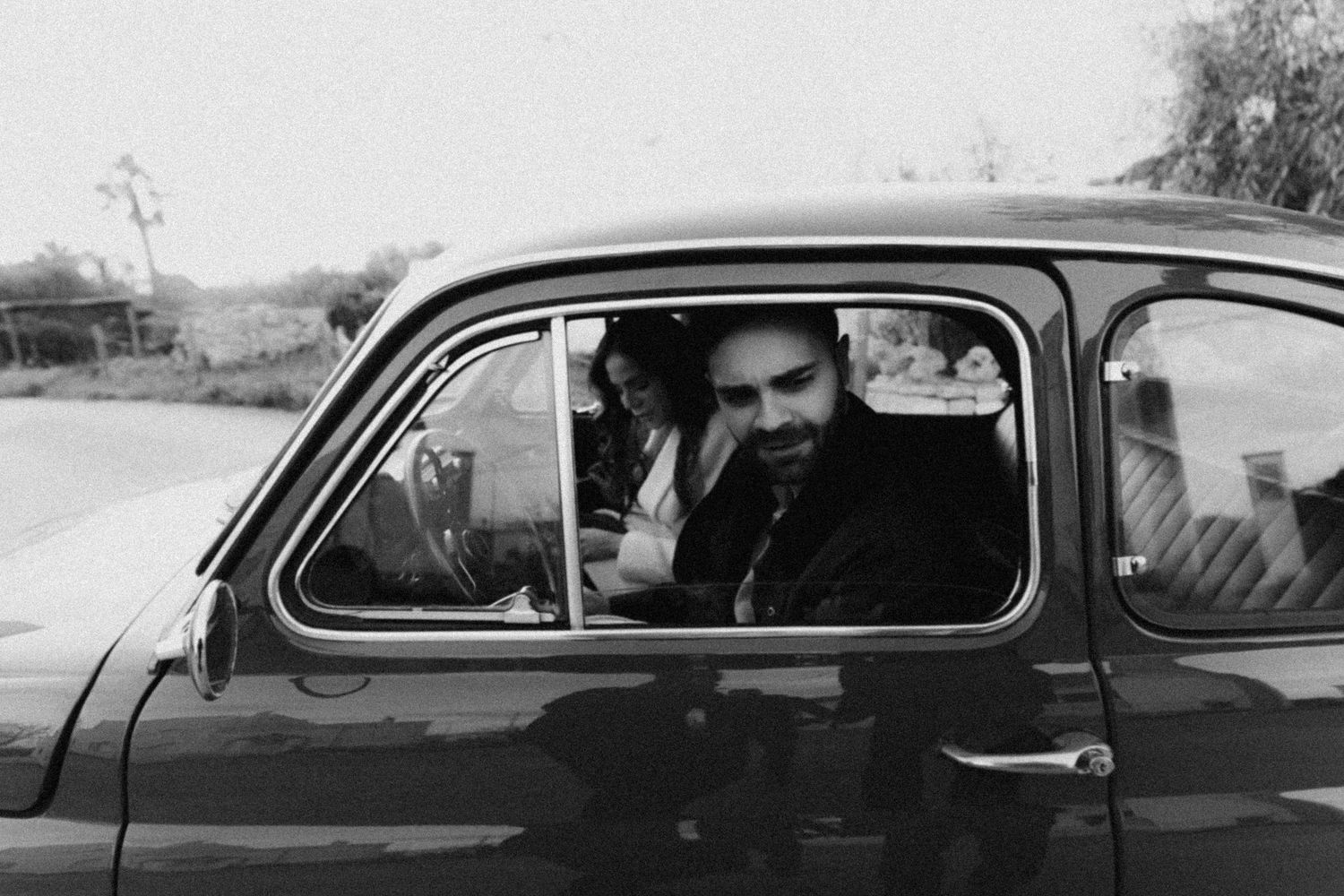 Black and white photograph of two people seated in a vintage car on a rural road.