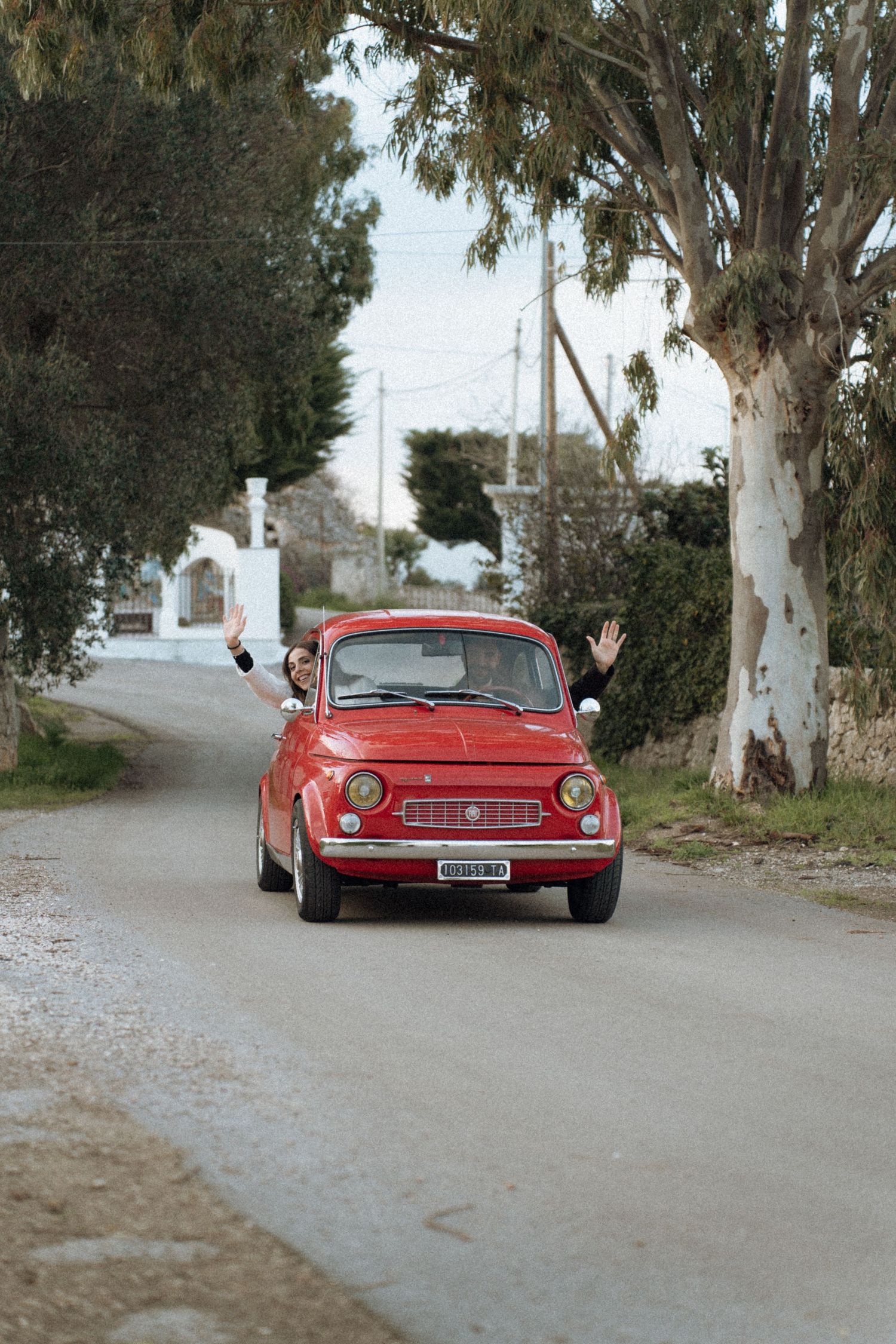 A red vintage Ferrari sports car parked on a gravel road lined with eucalyptus trees.
