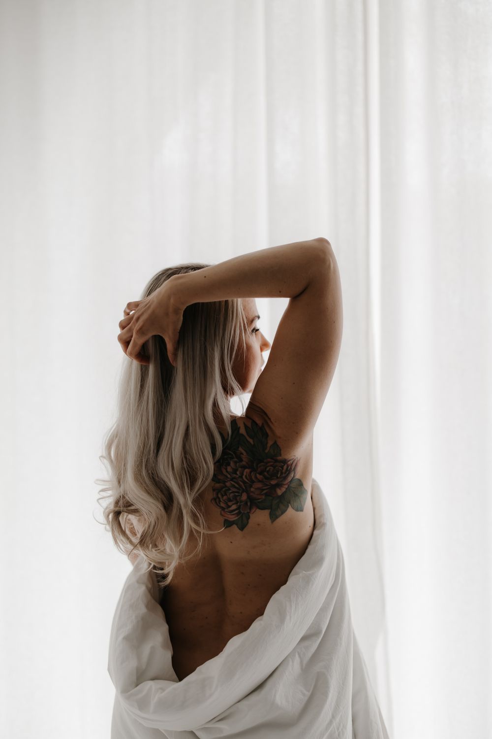 Blonde woman with floral back tattoo posing against white curtains with hands raised.