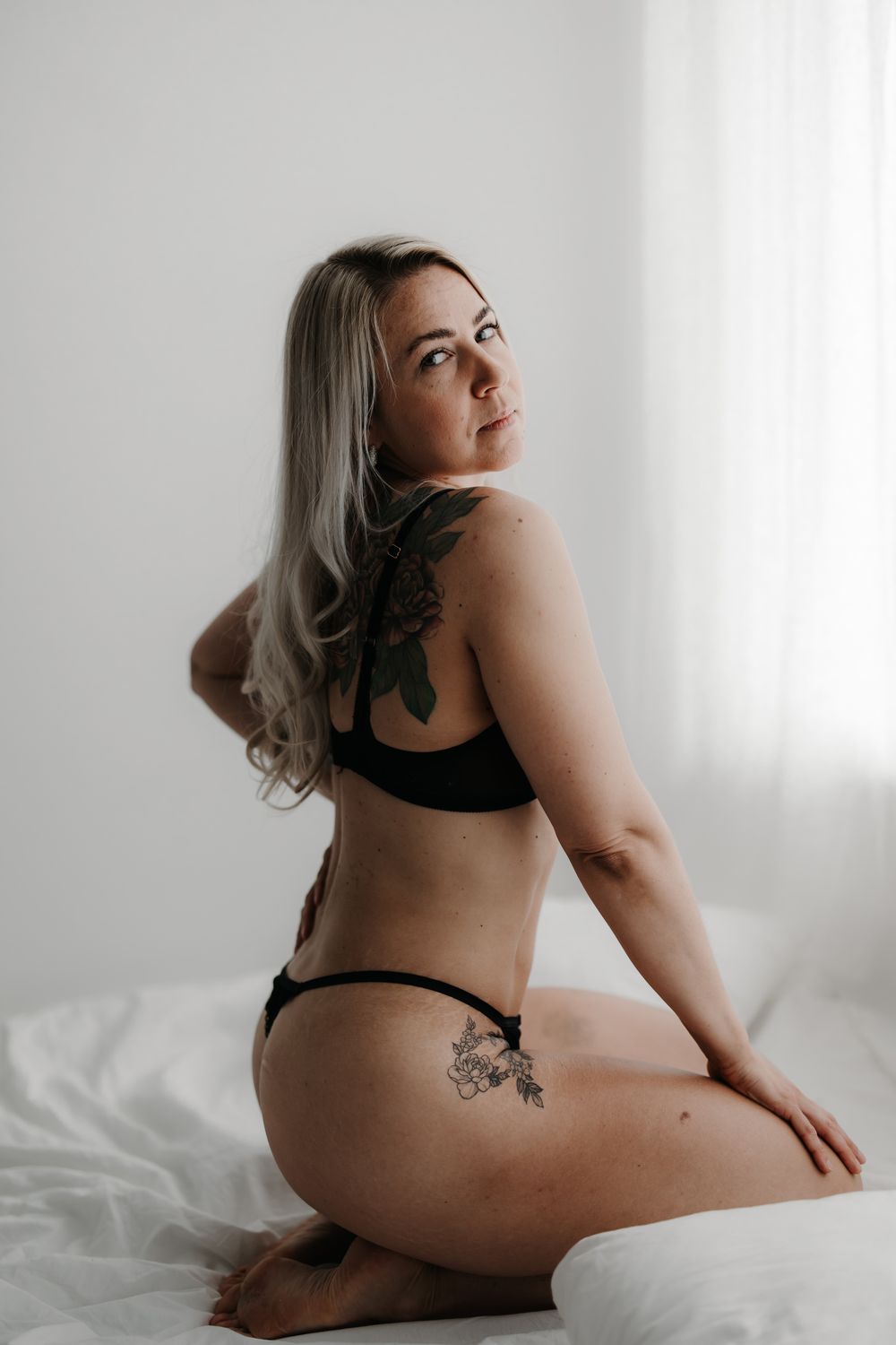 Woman in black lingerie with floral back tattoo sitting on white bed, looking over shoulder.