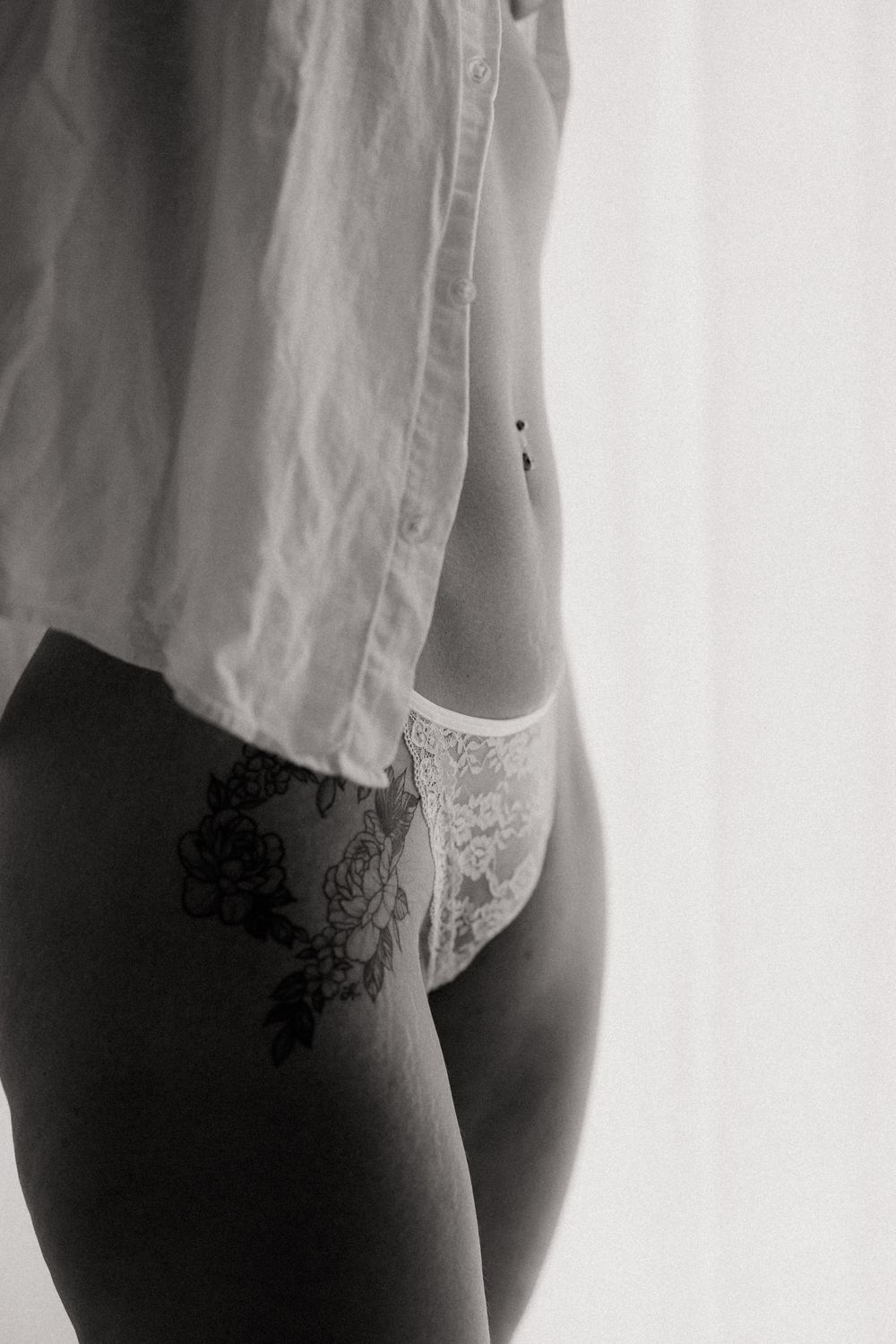 Black and white photo of a woman in lace underwear and open shirt, featuring a floral tattoo on her hip.