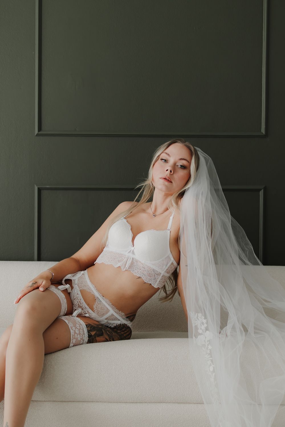 Bride in white lingerie and veil sitting elegantly on a cream sofa against a dark green wall.