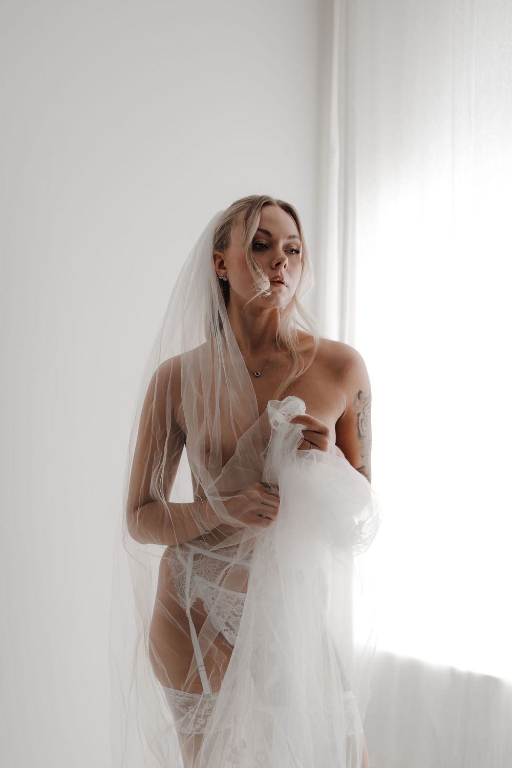 Bride in white lace lingerie and veil poses elegantly near a bright window in a boudoir photography session.