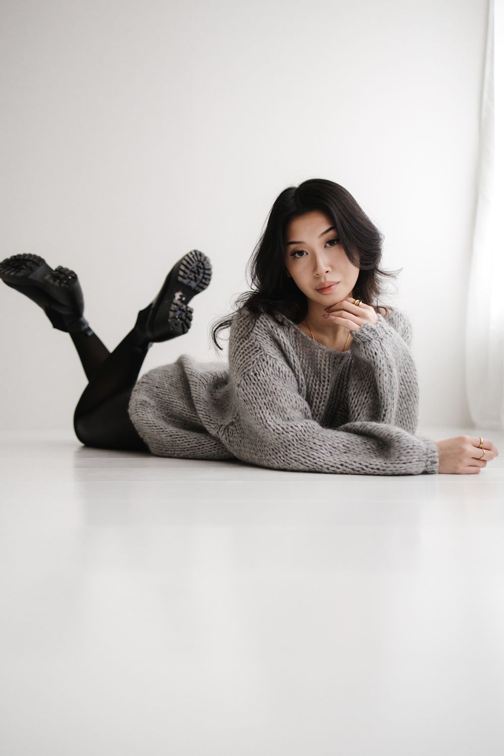 Fashion model in cozy gray sweater poses lying down against white backdrop in minimalist photo series.