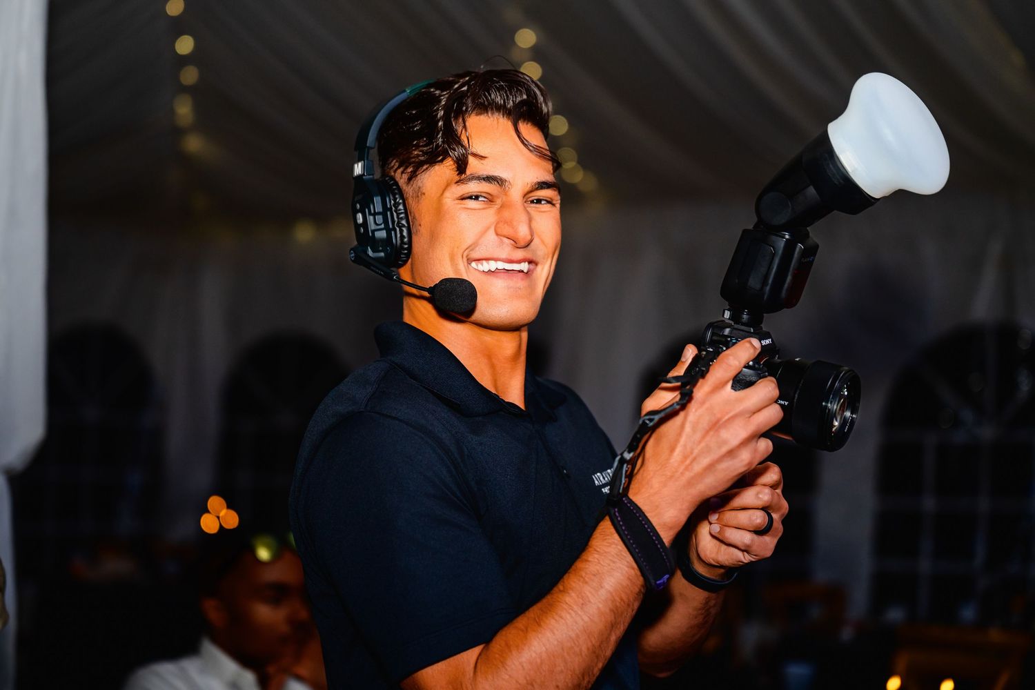 Professional photographer holding a camera outdoors with a happy expression amidst greenery and flowers.