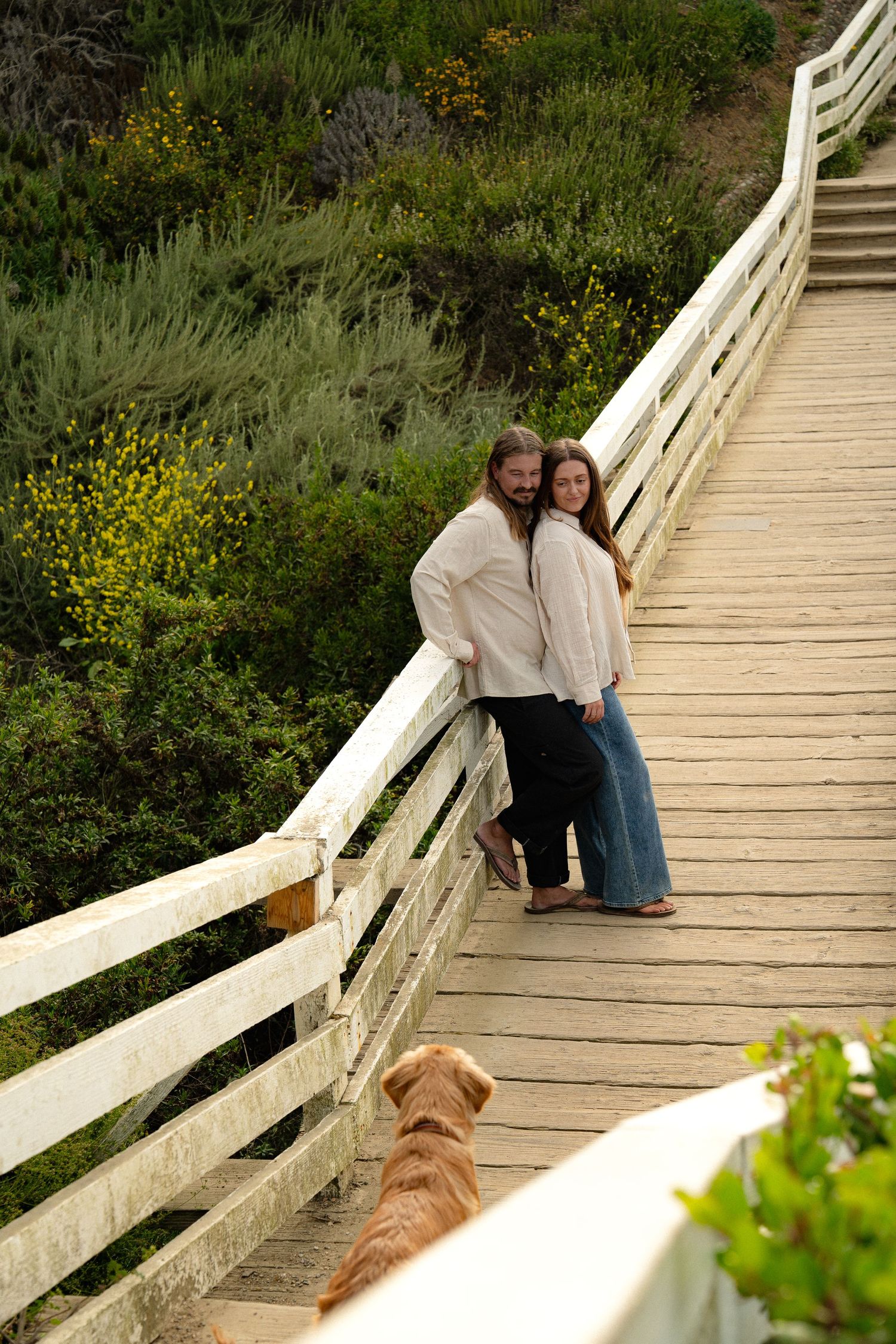 A couple and their dog share a moment on a wooden boardwalk surrounded by coastal vegetation.
