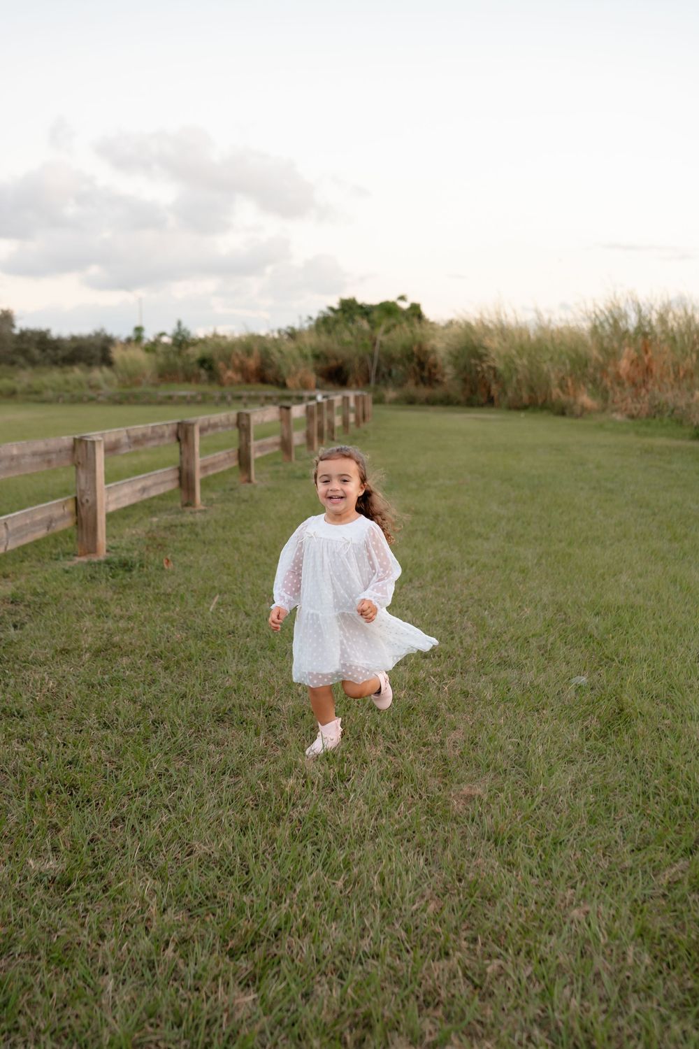 A small child in a white dress runs playfully along a wooden fence in a grassy field.