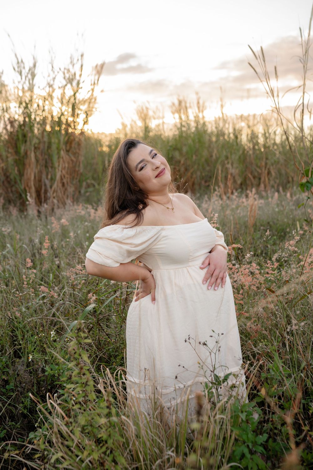 A white dress flows in a golden field at sunset as someone poses gracefully among tall grasses.