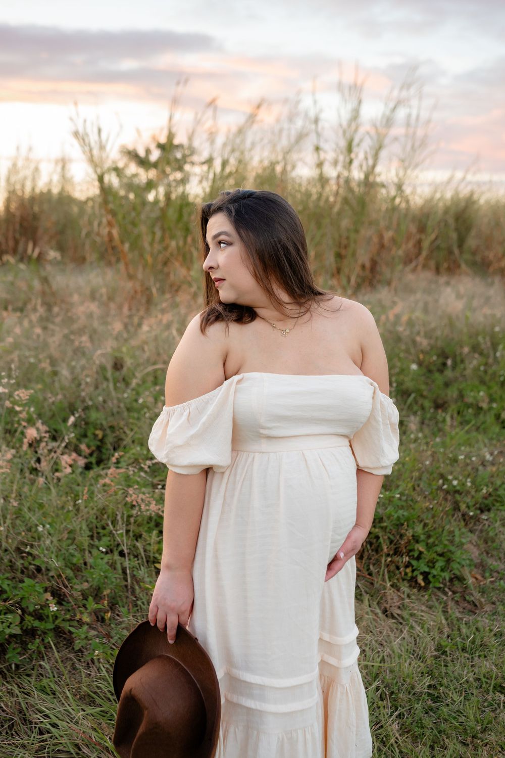 A person in a flowing white off-shoulder dress poses gracefully in a field at sunset holding a brown hat.