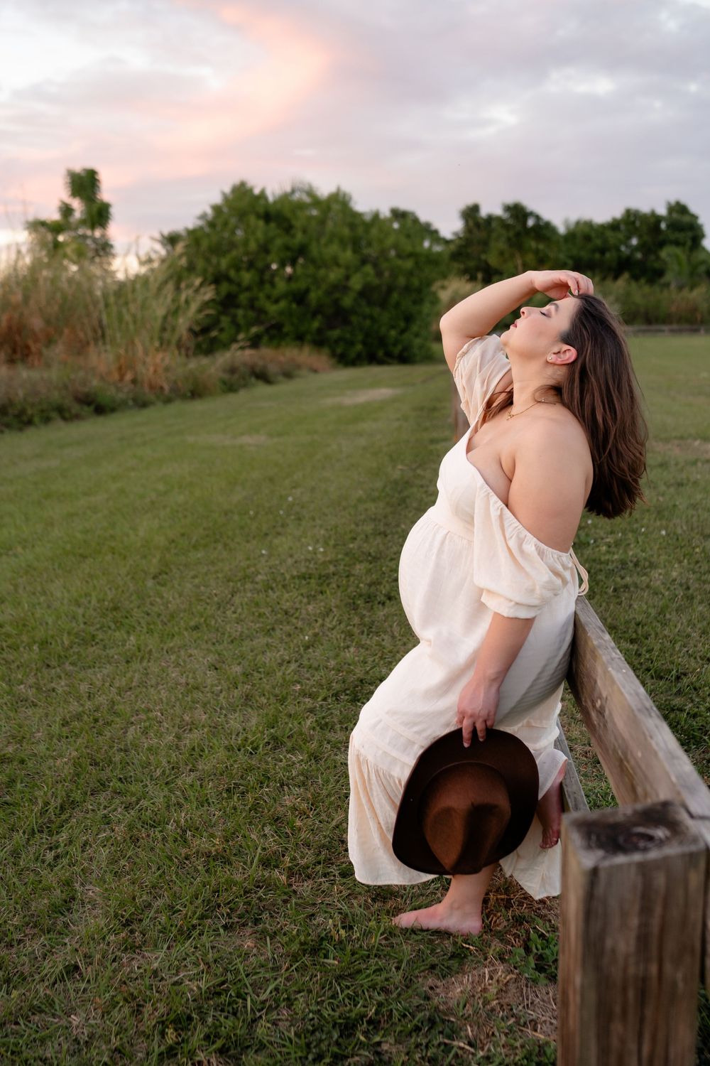 Ethereal maternity photo of a woman in a flowing white dress holding a brown hat at sunset in a grassy field.