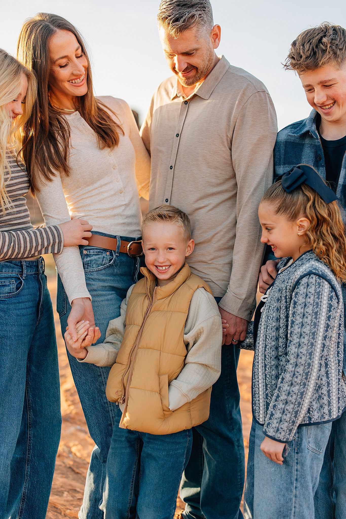 A family wearing casual clothing stands together in a row during golden hour, sharing laughs and embraces.