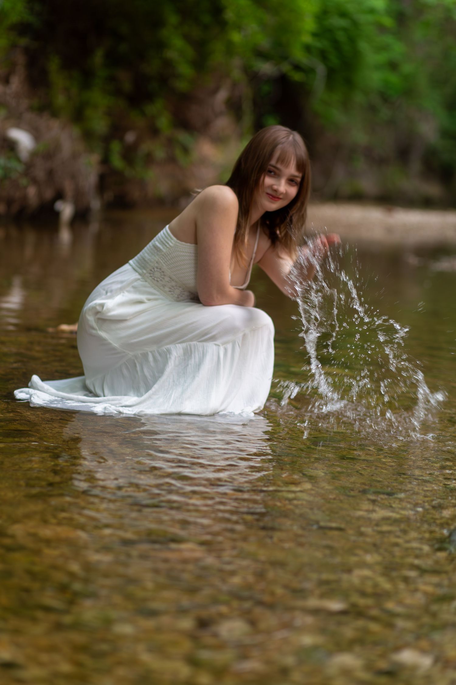 A person in a flowing white dress crouches in shallow water, creating splashes while posing in a natural stream setting.