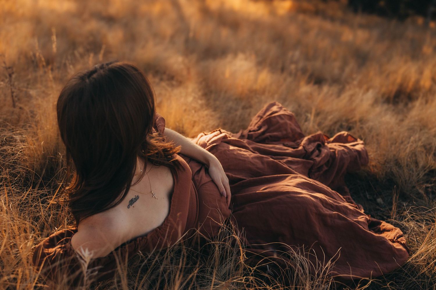 A figure in a flowing burgundy dress lies gracefully in golden autumn grass during sunset.