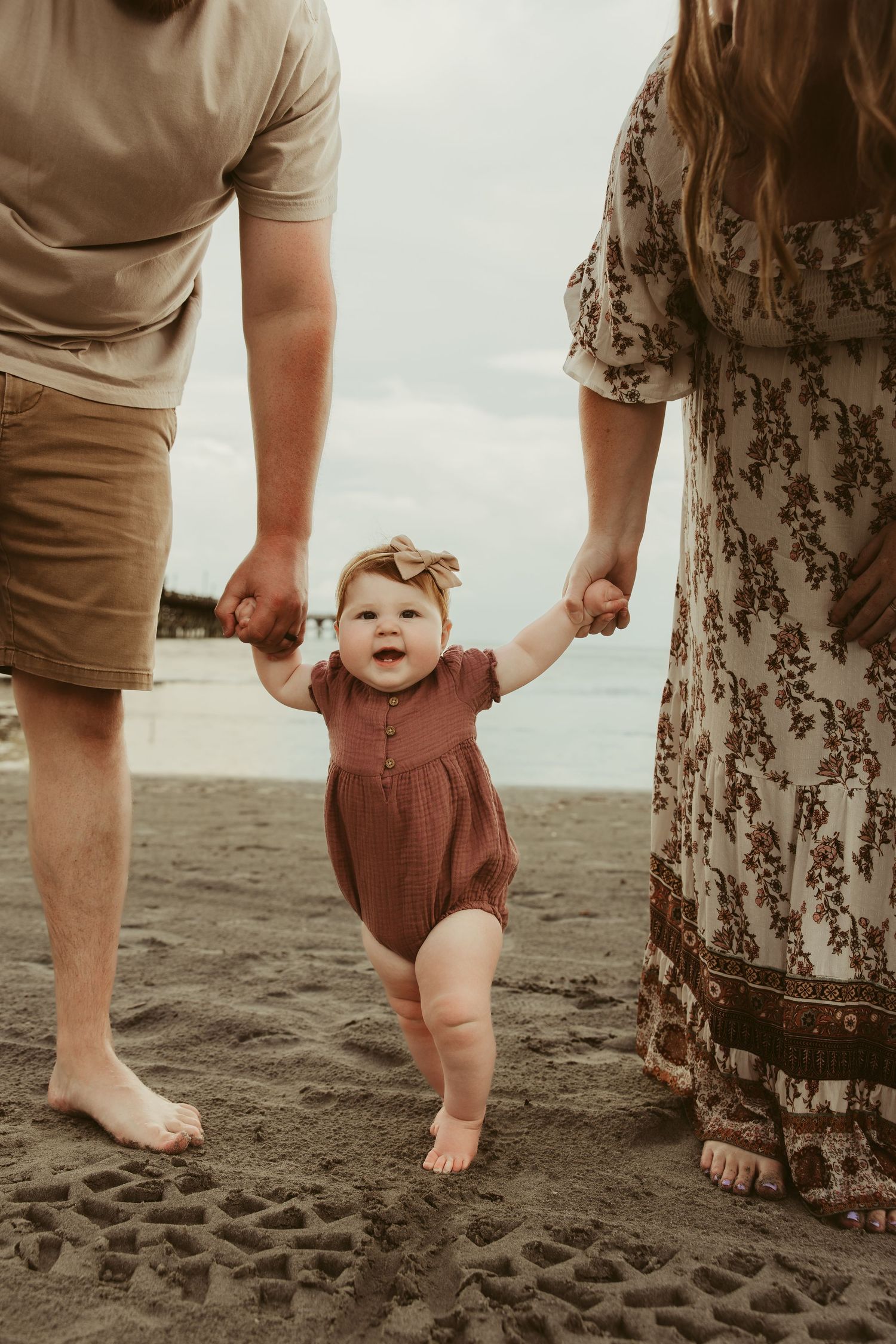 Baby girl taking first steps on beach held by parents hands.