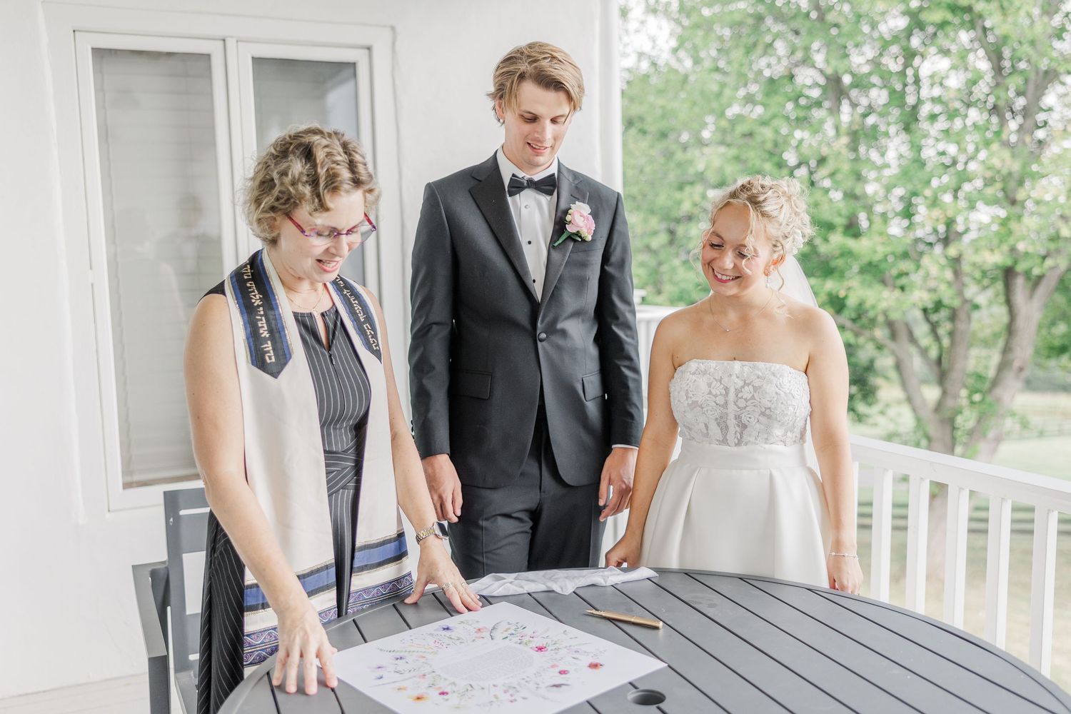 Three people examine paperwork on a gray table on an outdoor porch.