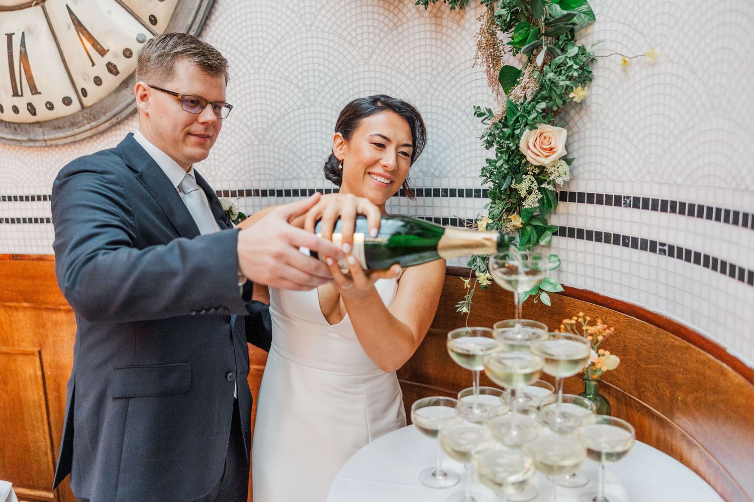 Two people pour champagne into a tower of glasses at an elegant celebration with floral decor and vintage clock.