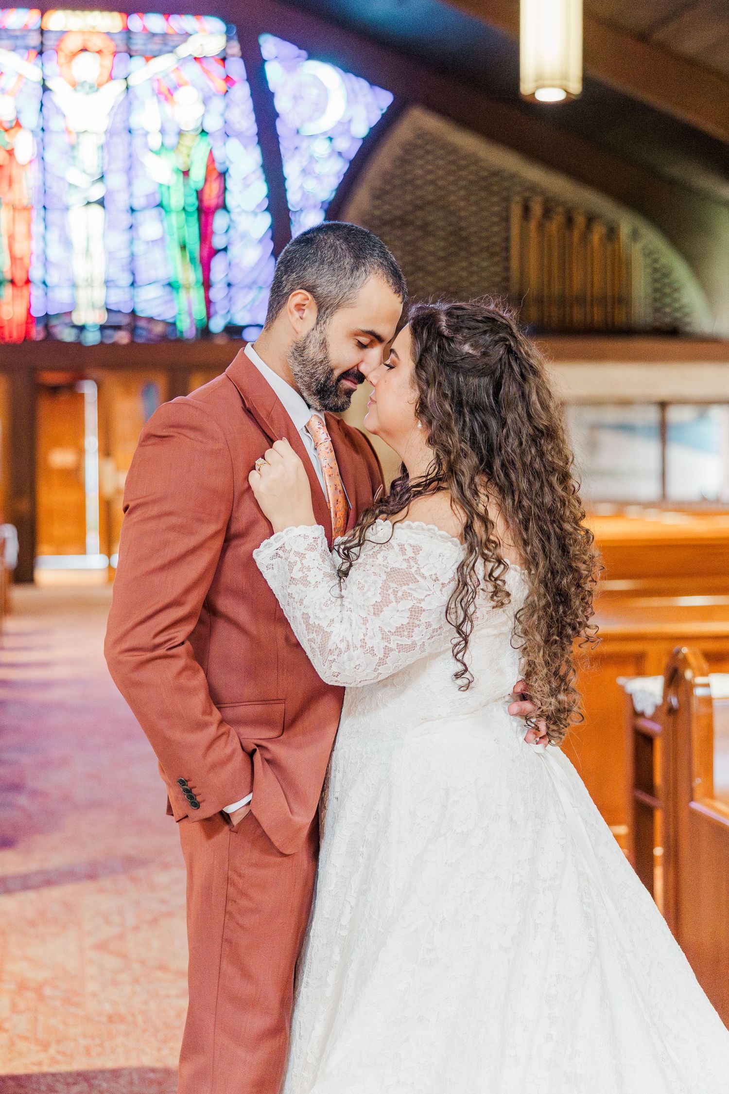A romantic wedding couple embraces in a church with stained glass windows and wooden pews in the background.