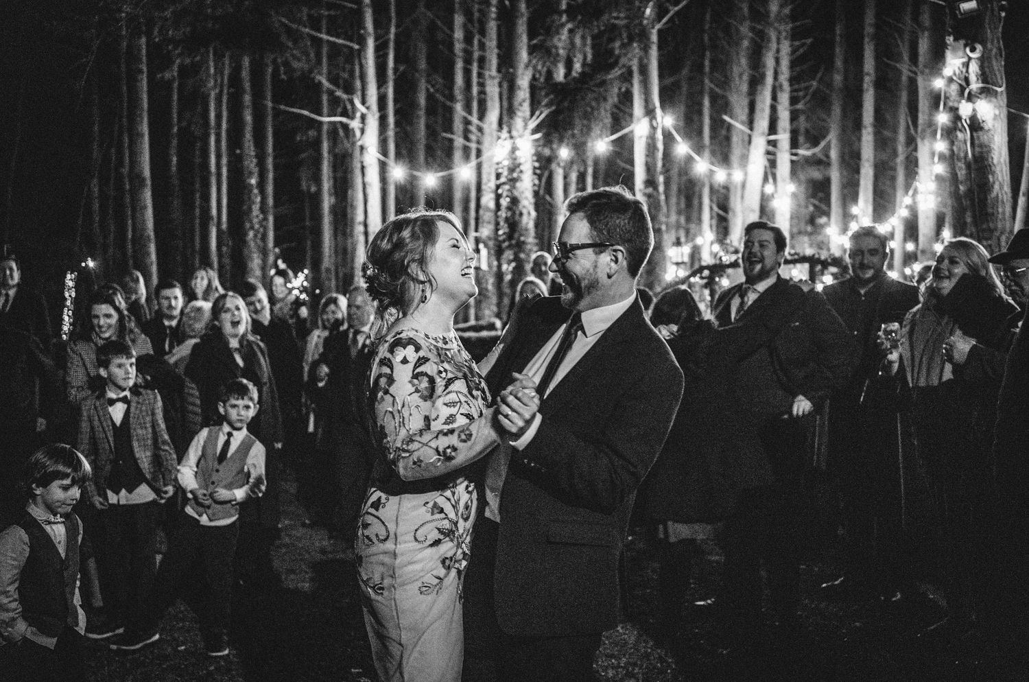 A wedding party dances and celebrates during a reception in a dimly lit venue with string lights overhead.