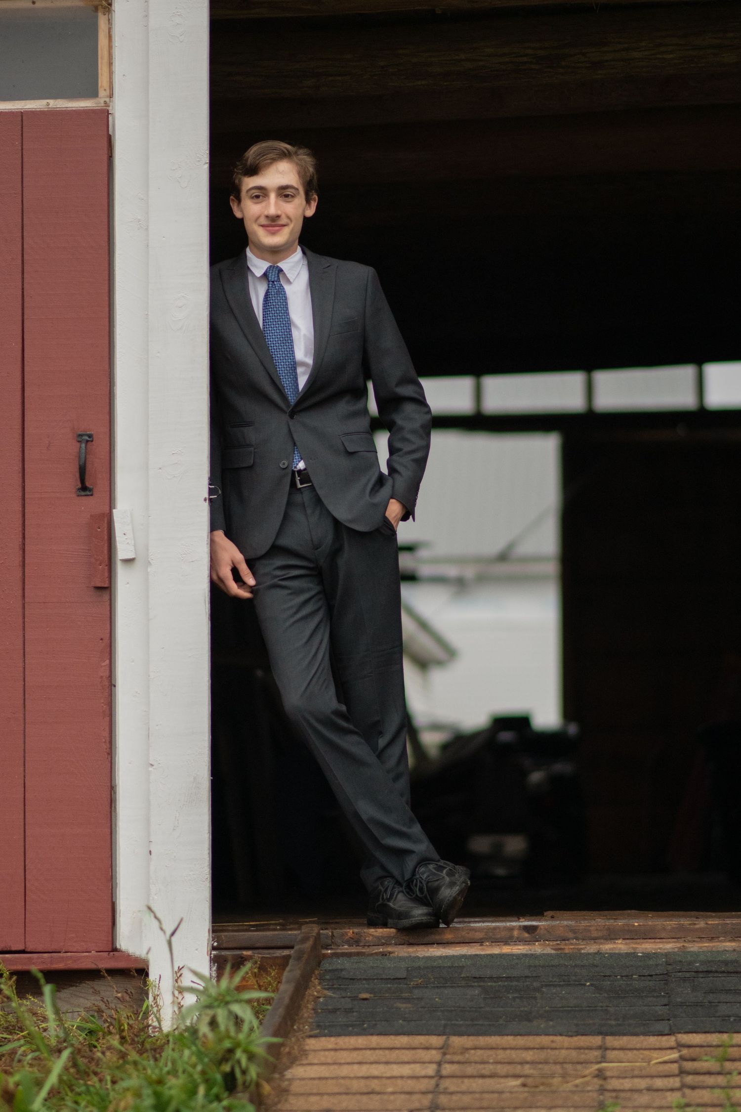 Confident young man in gray suit and blue tie leaning against barn doorway smiling. Photo by Xenophon Photography in Maine.