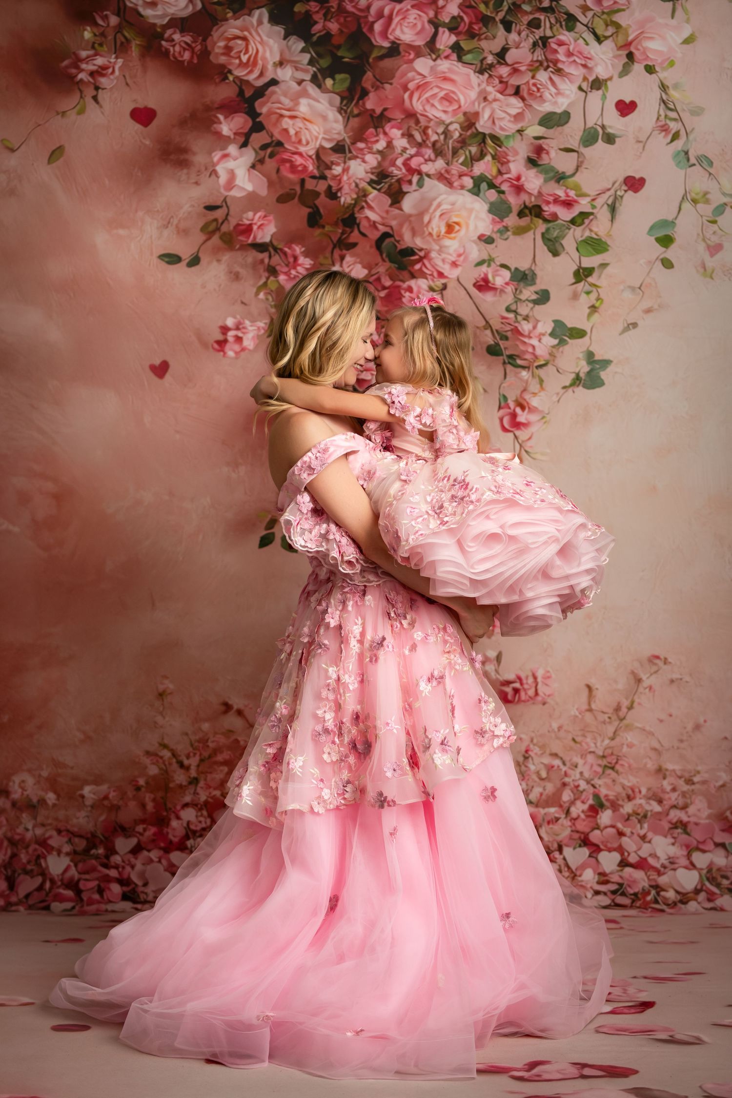 Mother and daughter in matching pink gowns pose together in a dreamy floral studio setting.