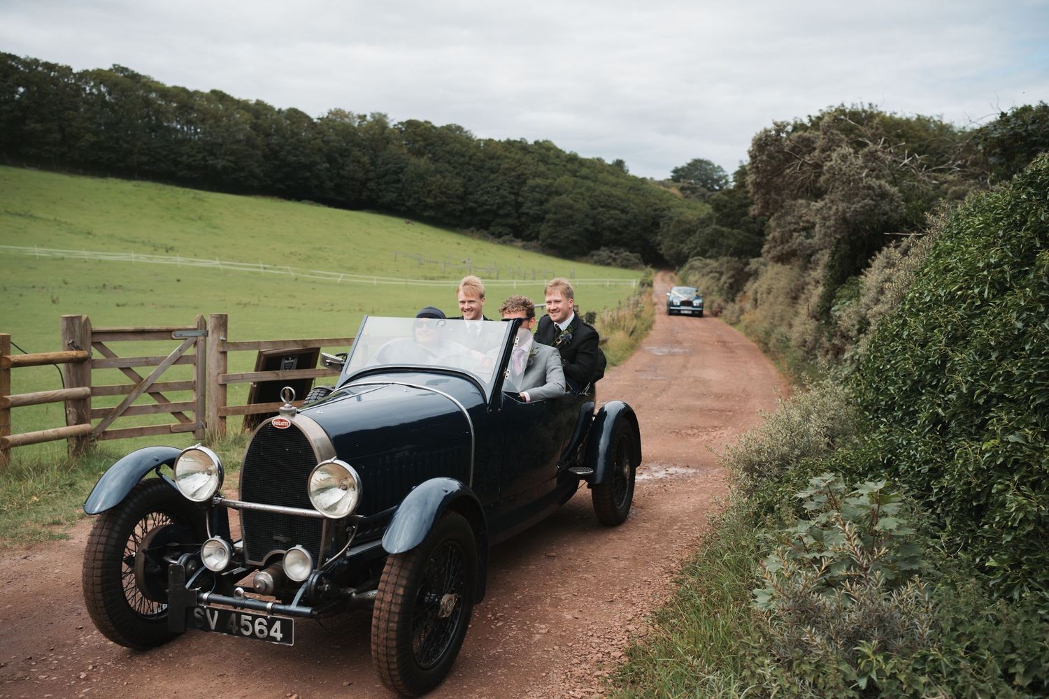Vintage classic cars driving along a scenic countryside dirt road on a sunny day.