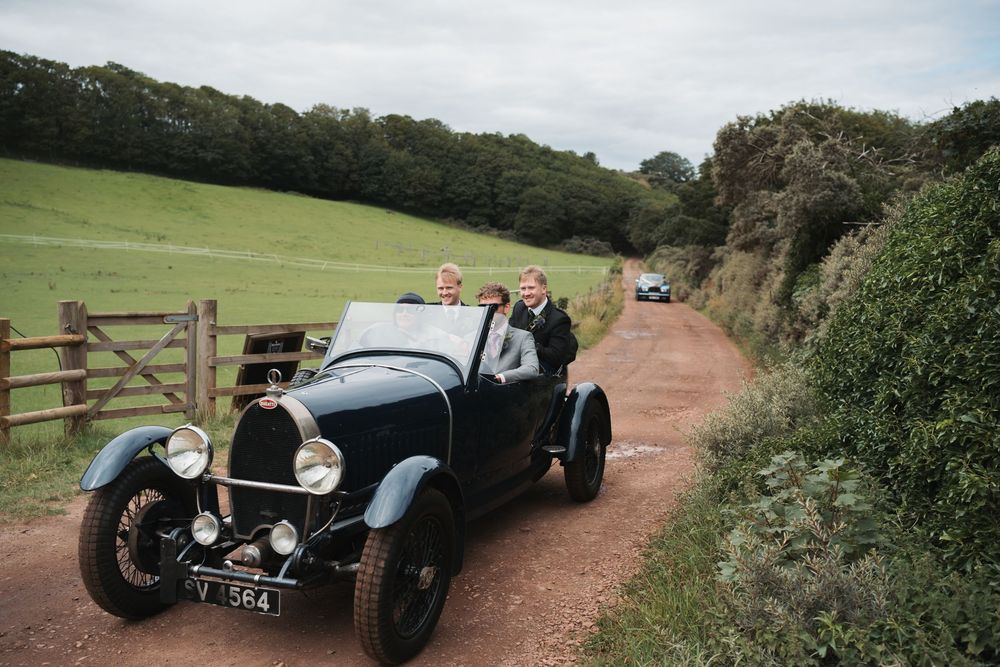 Vintage classic cars driving along a scenic countryside dirt road on a sunny day.