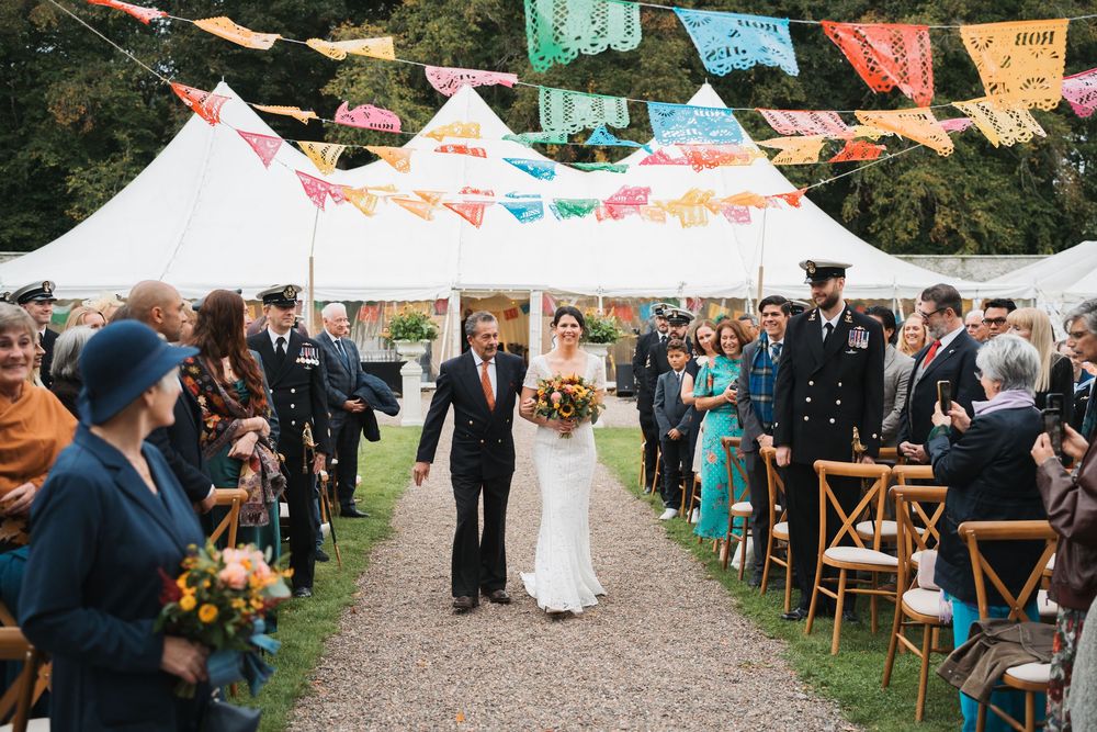 Colorful papel picado banners hang above an outdoor wedding celebration with guests seated at long tables under a tent.