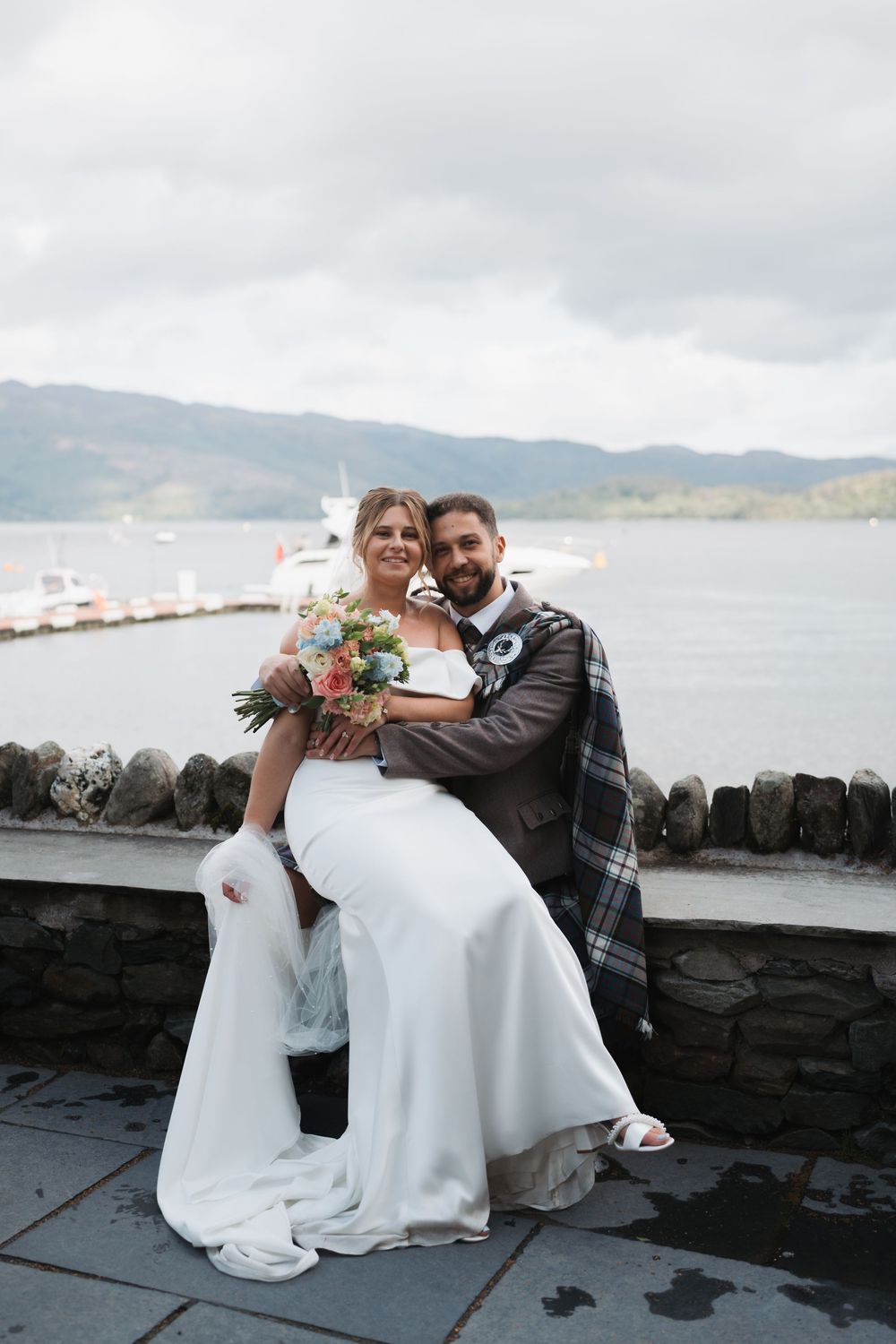 Newlyweds share a joyful moment sitting by a stone wall overlooking a scenic waterfront.