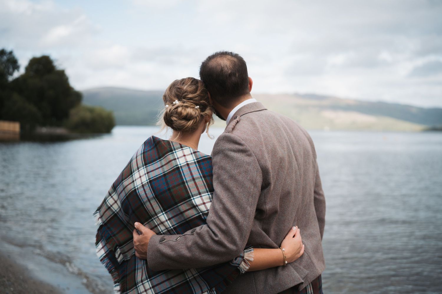 Couple embraces while standing near a scenic lake with mountains in the background.