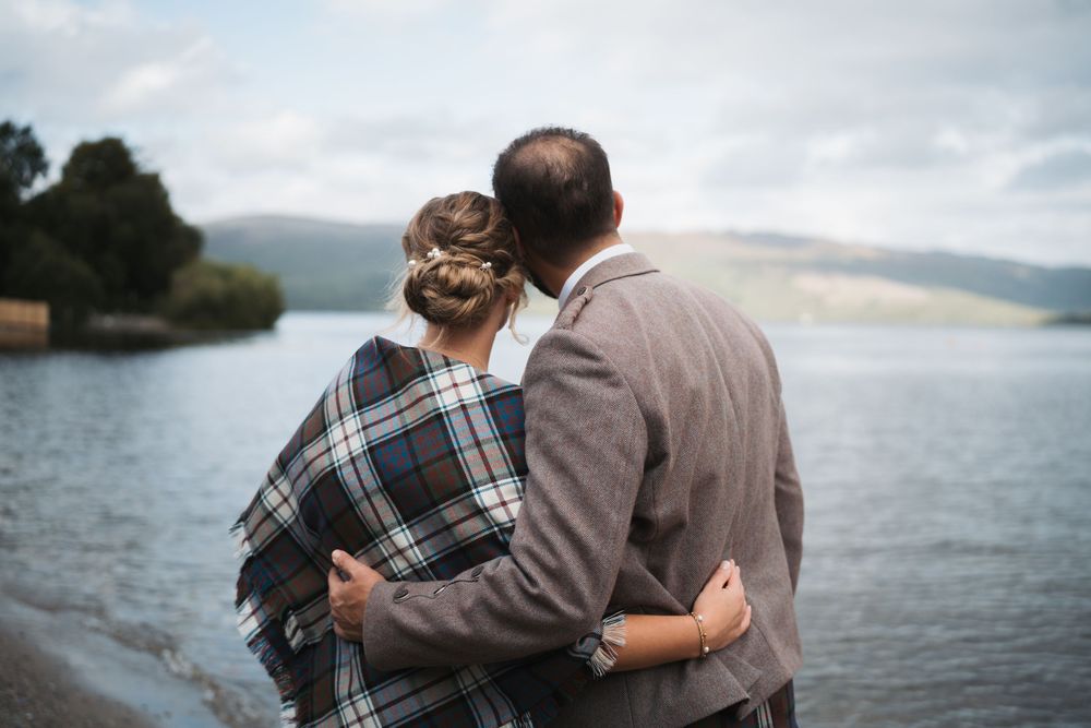 Couple embraces while standing near a scenic lake with mountains in the background.