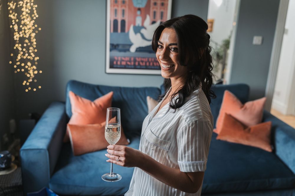 Woman in white sweater relaxes on blue couch with champagne glass while taking selfies in cozy room with lights.
