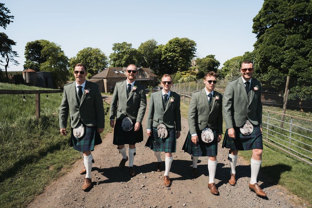 A row of groomsmen in matching sage green suits walk down a dirt path in a rustic outdoor setting.