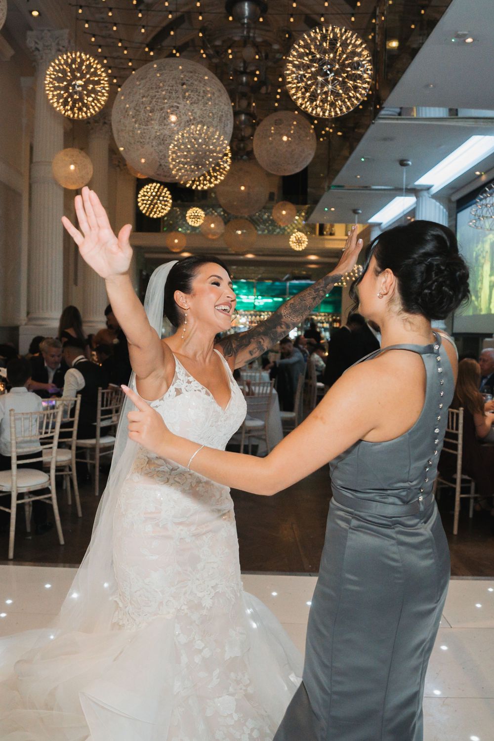 Two people in formal attire dancing joyfully under decorative hanging lights at an elegant event venue.