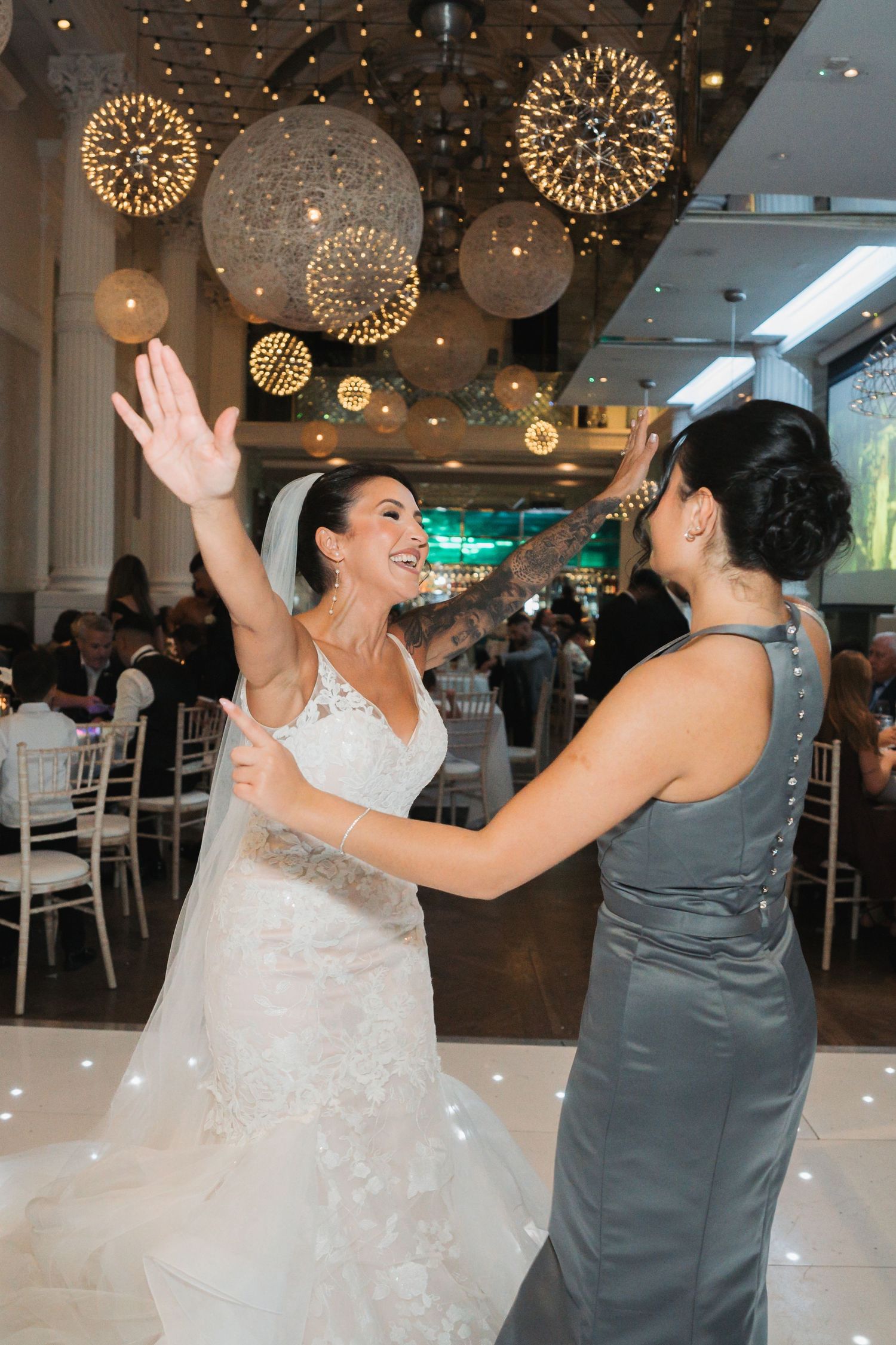 Two people in formal attire dancing joyfully under decorative hanging lights at an elegant event venue.