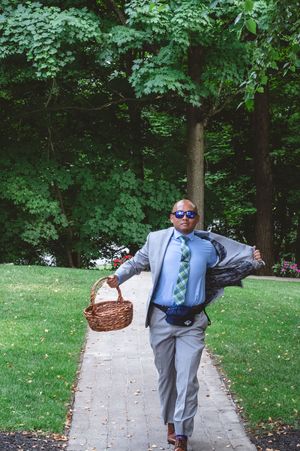 A person in a blue suit and tie walks along a stone path through a lush green garden while carrying a woven basket.