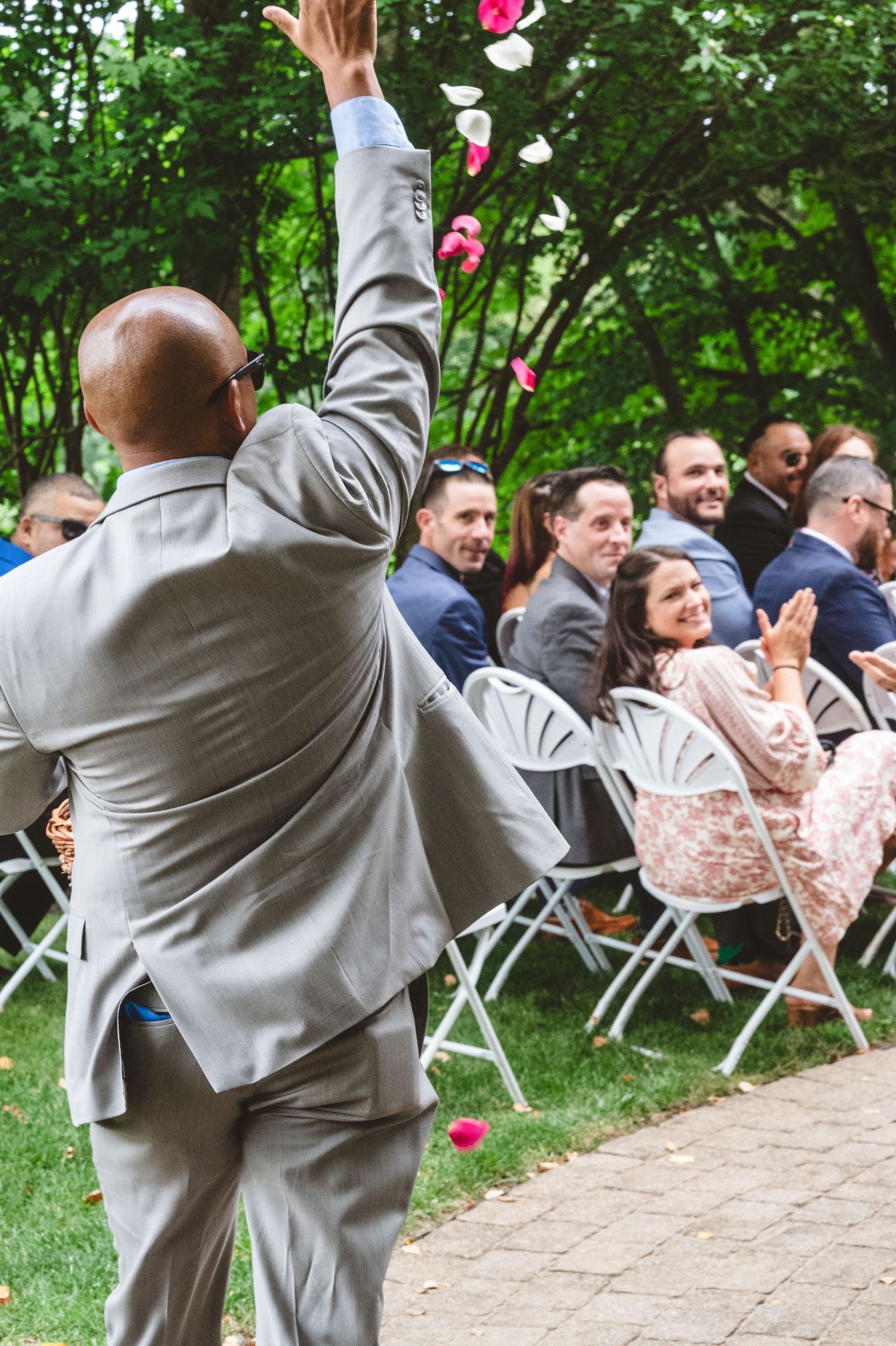 Groomsman in gray suit playfully kicks rose petals into the air during outdoor wedding ceremony.