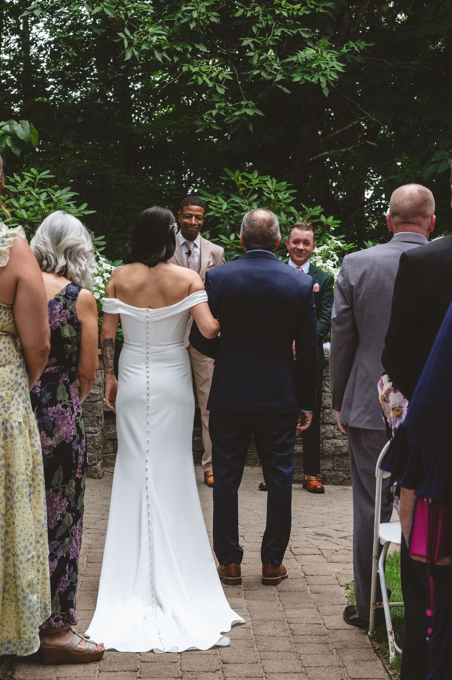 Wedding guests gather on a stone patio surrounded by lush greenery during an outdoor ceremony.