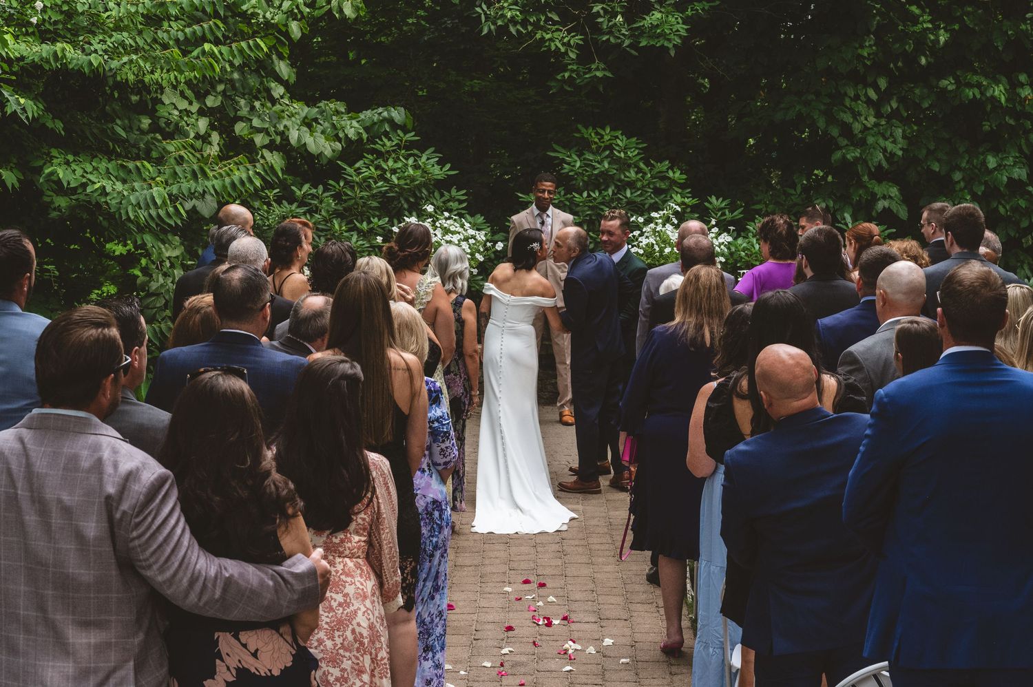 Wedding guests gather outdoors under green trees for an intimate garden ceremony.