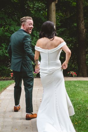 A couple in wedding attire, with a green suit and white dress, walking and laughing together on a garden path.