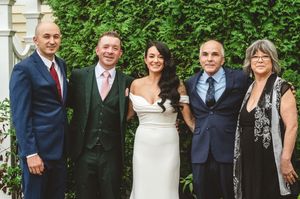 Wedding party poses together outdoors against lush green foliage and stone walls.
