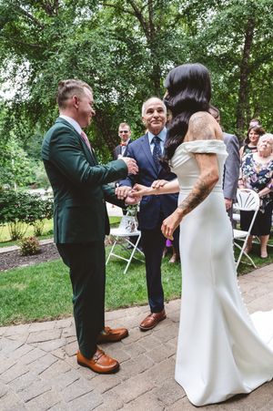 Wedding party members dance and celebrate on a stone patio surrounded by trees.