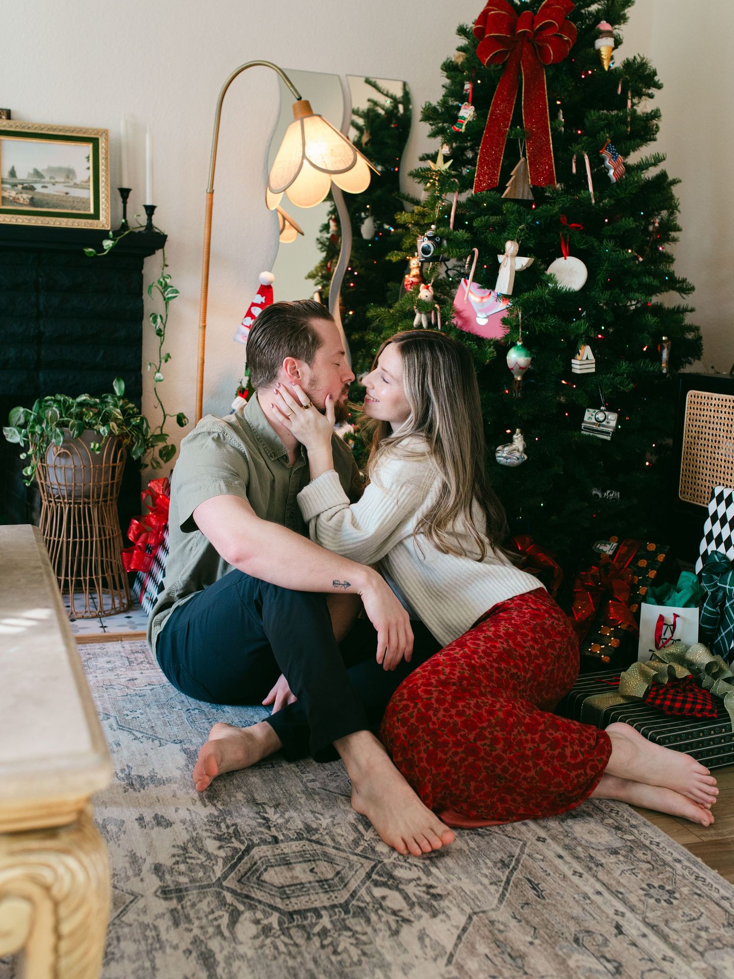 Couple sits together on floor by decorated Christmas tree sharing intimate moments in series of romantic holiday photos.