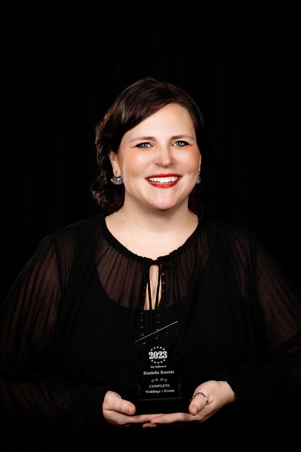A person in black clothing smiles while holding an award trophy against a dark background.