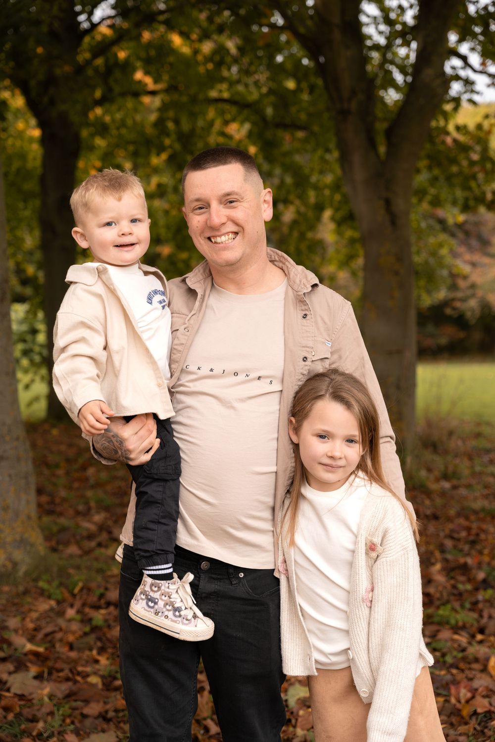 Family wearing coordinated beige and navy outfits poses together among autumn trees and fallen leaves.