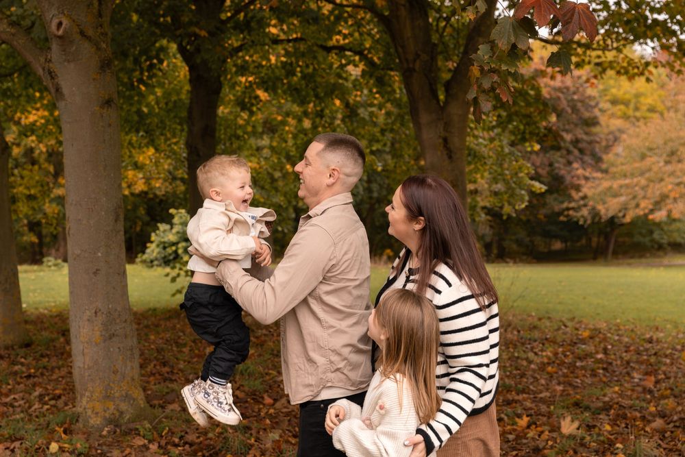 A family enjoys a playful autumn moment together in a park with fallen leaves and golden trees in the background.