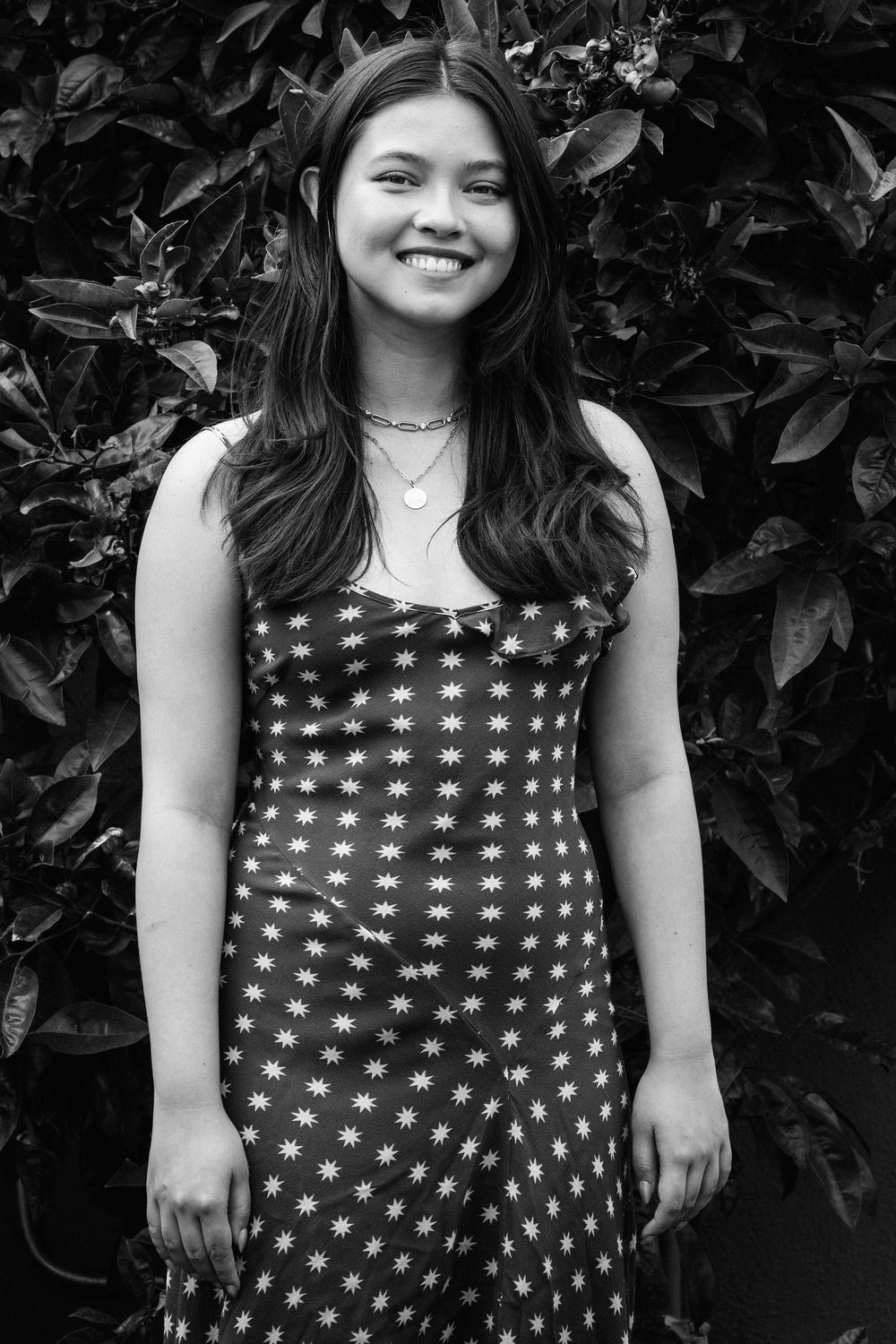 Black and white portrait of a person wearing a star-patterned dress standing in front of lush foliage.